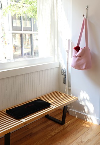 A vertical image shows the two works, Lap Pad (no regrets) and Sensory Bag (pink and red), installed together. The soft black lap pad with the red embroidered text “NO REGRETS” rests on a wood bench to the left underneath a window that lets in sunlight. O