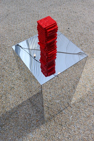 Photographed from above, we see the mirror pedestal blend into the sandy tan color ground of tiny pebbles, the ground is made sturdy by a hard to see mat of small hexagons that hold the pebbles. The red Covid rapid test Jenga tower is reflected downwards on the mirror pedestal alongside string lighting and lines warped by the mirror from an overhead structure.