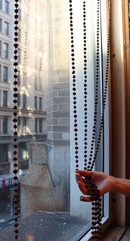  5 chains of roasted coffee beans, distanced about 8 inches apart, hang like a beaded curtain in front of a large vertical window that looks out on a large New York City building with a grid of windows. A hand in the bottom right of the image reaches tenderly to hold 4 of the coffee bean chains. The chains continue on upwards beyond the image illuminated by a golden sunlight. 