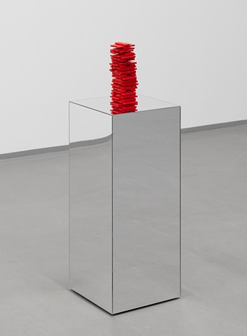 Dozens of Covid rapid tests are painted bright red and stacked like a Jenga tower. The tests are rectangular, a few inches long and about a quarter inch thick. The tower is roughly 8 inches tall and is arranged mid-game so there are tests missing here and there among the rows of 3. It rests on a pedestal made of mirrors on every side which reflects the red tower downwards. We see the pedestal from a three fourths angle in front of a white wall and on gray concrete floor which are reflected on the sides of the pedestal.