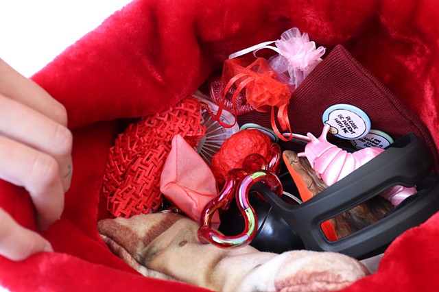 A hand out of focus pulls open the bag to reveal various items nestled together in the fuzzy red lining of the sensory bag. Most items are also pink and red like the bag. All the visible items are described in the caption below.