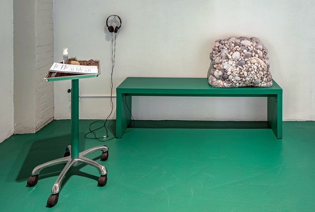 A green bench is sits on a green floor, and a rolling metal tray is placed in front of it with its shaft also painted in the same cool toned green. On the bench is a large lumpy fleece pillow that is printed with an image of a large pile of small rocks. T
