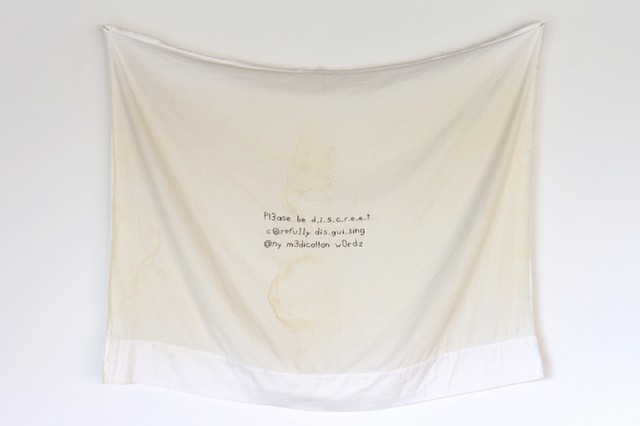 Image Description: A sweat stained pillowcase hangs on a white wall. The fabric dips in the middle creating V shaped folds. It has been split open and appears as a continuous sheet of fabric. In the center of the fabric is small text stitched in black thr