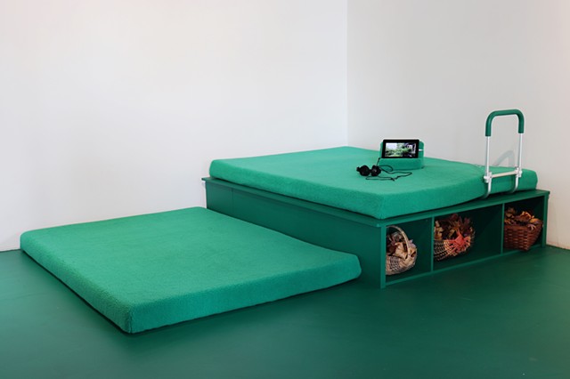 A bed frame is installed in the corner of a gallery with white walls. The floor is painted "OSHA Safety Green", and the bed frame is painted in the same color. On top of it is a memory foam mattress topper covered in emerald green sherpa fleece. A second 