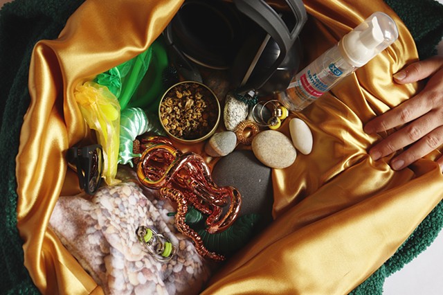 A light skinned hand on the right of the image opens the sensory bag showing its contents and shimmery gold satin lining. Huddled together are various sizes of rocks, stretchy stim toys in mesh green and yellow bags, green and gold acupressure rings, an o