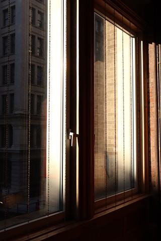 Shot at an angle, two large vertical windows surrounded by darkness let in warm golden light which brings a beaded curtain of 10 chains of coffee beans into focus, 5 on each window distanced about 8 inches apart.  Behind the coffee chains through the window on the left is a tall beige older New York City building with a grid of many windows.