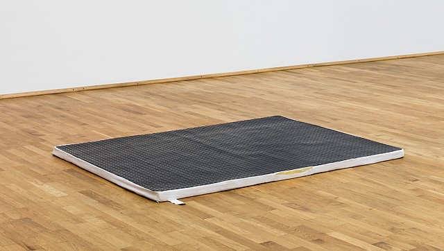 Resting in the middle of a wooden floor and in front of a white museum wall, a mattress topper with a white fabric cover can be seen from underneath black diamond plate rubber flooring which has been cut to fit the dimensions of the mattress topper. Light shines on the textured rubber surface. The yellow foam of the mattress topper peaks out the from a tear in the white cover and a small tag extends outwards from the fabric. 