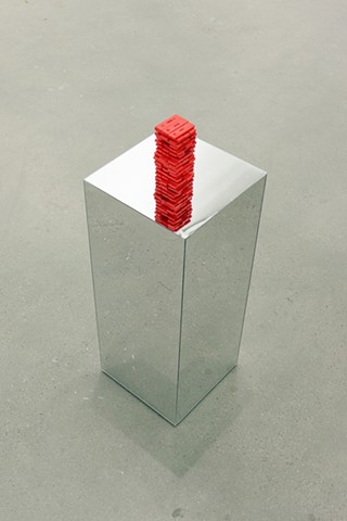 Dozens of rapid home Covid tests are painted bright red and stacked like a Jenga tower. The tower is arranged mid-game so there are tests missing here and there among the rows of 3. It rests on a pedestal made of mirrors on every side which reflects the red tower downwards. We see the pedestal from a three fourths angle from slightly above on gray concrete floor which is reflected on the sides of the pedestal.