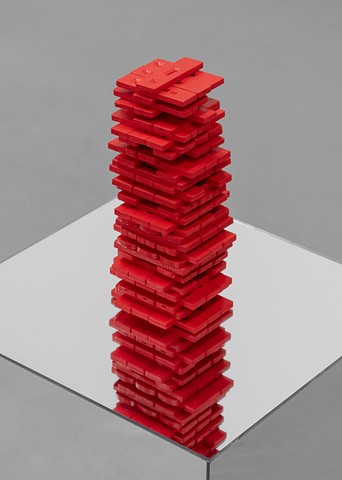 Dozens of Covid rapid tests are painted bright red and stacked like a Jenga tower. The tests are rectangular, a few inches long and about a quarter inch thick. The tower is arranged mid-game so there are tests missing here and there among the rows of 3. It rests on a pedestal made of mirrors on every side. A close up of the tower shows it reflected downwards on, or seemingly into, the mirror surface. 