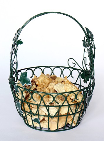 A dark green wire basket, with a metal vine of leaves climbing up both sides of the rounded handle, holds a few dozen small pieces of bumpy textured burl wood. The metal leaves on the basket have faded white edges. The basket has a wire pattern that look