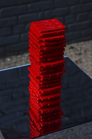 The sunset shines on a close up of the red covid rapid test stack, which is in rows of 3 like the game Jenga. The moody lighting creates high contrast between the bright red of the stack and the dark grey brick background reflected in the mirror under the stack of the mirror pedestal it sits on. The red stack is also reflected downwards. 