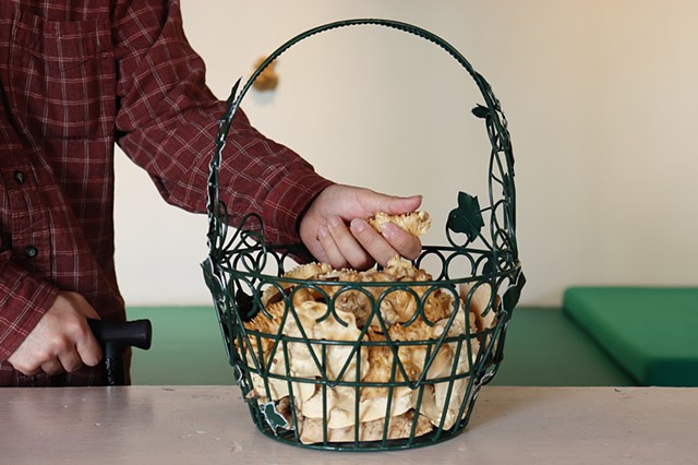 A dark green wire basket, with a metal vine of leaves climbing up both sides of the rounded handle, holds a few dozen small pieces of bumpy textured burl wood The metal leaves on the basket have faded white edges. The basket has a wire pattern that looks 