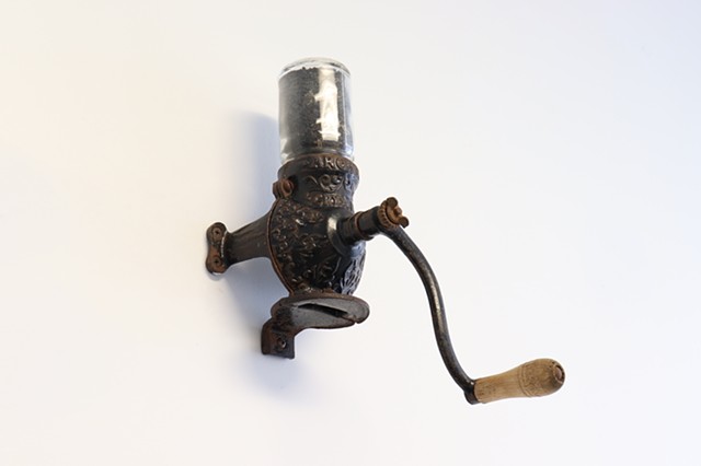  An antique coffee grinder is mounted on a white wall and seen at an angle from slightly below. It’s dark brown, covered in rust and has a rounded body with flanges protruding from the sides with matching rust color screws. A curved crank with a wood handle extends from the center of the coffee grinder. Closer look shows ornate details and the words “Arcade Crystal”. A glass jar about the same size as the body of the grinder is attached upside down to the top of the grinder. Inside are dark gray ashes with bits of debris.