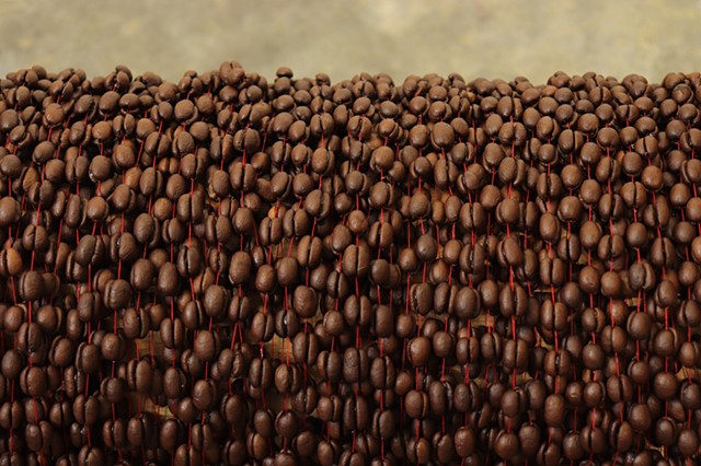 A close up of the center of a wooden cable reel, laying horizontally, is fully covered by layers of countless coffee beans, which have been glued together in pairs on a continuous red thread like beads on a necklace. 