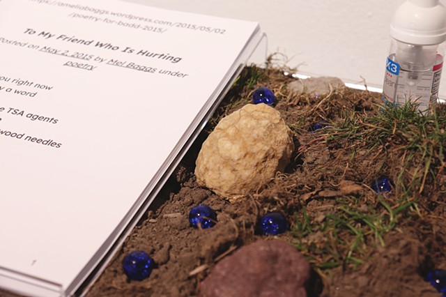 A close up of a metal tray filled with dirt and bits of grass. In the center of the image is a tan color geode that has not been cracked open. Around it are a couple other rocks, deep blue marbles, a small bottle of hand sanitizer, and an acrylic tray that holds a transcript of a poem, |https://ameliabaggs.wordpress.com/2015/05/02/poetry-for-badd-2015/|To My Friend Who is Hurting| by Mel Baggs.
