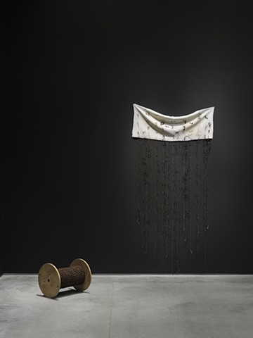  An off-white pillowcase with a swirly texture has been split open revealing a doubling of stains from where a head might rest. It’s hung high and horizontally on a black  wall and spot lit. The fabric dips slightly in the middle. Cascading from the fabri