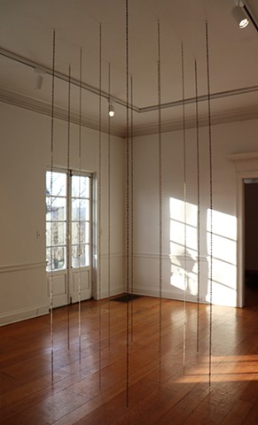 Chains of coffee beans are huddled in the center of a gallery with a wood floor and white walls.  The chains are hung in an inner circle of 6 unroasted coffee bean chains, and an outer circle of 6 roasted coffee bean chains, with enough space to stand in the middle. In between the chains of the inner circle is about a foot and a half of space, and it is about the same distance between the inner chains to the outer circle which is spread farther apart. Warm sunlight shines into the space from a door behind the installation.