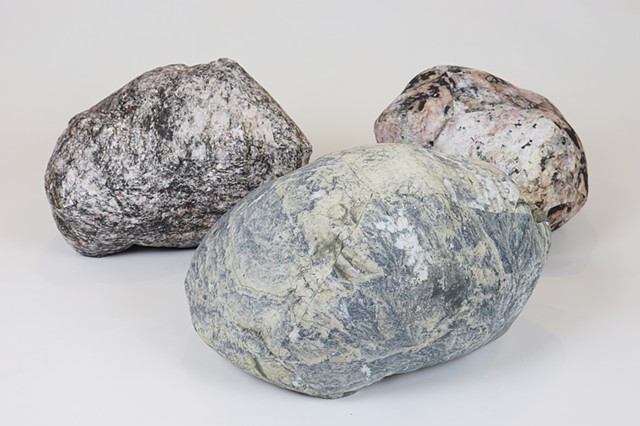  A family photo of three rock pillows with two positioned in the back and one in the center front against a white backdrop. Each rock has unique colors, shapes and textures, with rounded and more pointed sides. The front rock is primarily blueish light gray, the back left is primarily dark gray, and the back right is primarily light pink with black. Speckled and swirling granite and galaxy-like textures originating from close up photos of rocks are printed on cotton fabric creating a realistic appearance similar to a large rock you might find on the shorelines to stave off erosion. 