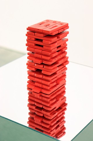 Dozens of rapid home Covid tests are painted bright red and stacked like a Jenga tower. The tower is arranged mid-game so there are tests missing here and there among the rows of 3. It rests on a pedestal made of mirrors on every side. Here we see a close up of the red tower that is reflected downwards in the mirror that it sits on. 