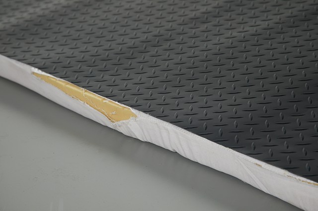 On a grey floor, yellow memory foam peaks through a tear in the seam of a white fabric mattress topper cover. The edges are frayed. Black diamond plate rubber flooring lies flat on top of the fabric covered foam.