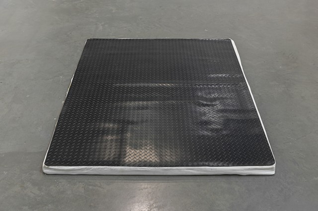 At Work (In Protest and in Care) lies on a dark gray museum floor and is shot straight on and centered in the image. The used memory foam mattress topper is covered by a white fabric cover and placed underneath black diamond plate rubber flooring which ha