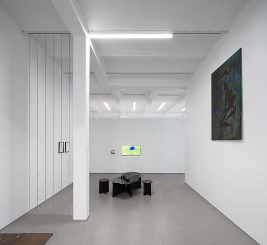 A view of the exhibition down the gallery hallway, with very tall white walls and a gray floor, shows an unroasted coffee bean curtain to the left of the image between the wall and a pillar. It is installed among other works including a video on a monitor and a black tactile curving table centered by the back wall, small framed works behind the coffee curtain to the left, and a large dark green painting hung right on the right wall. 