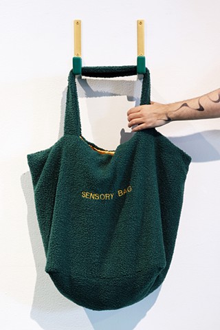 A light skinned hand with a forearm tattooed with an abstract curvy form reaches out from the right of the image to grab a bag hung on a white wall. The bag is a large saggy tote bag made of fuzzy dark green fabric and you can see a hint of the shiny gold