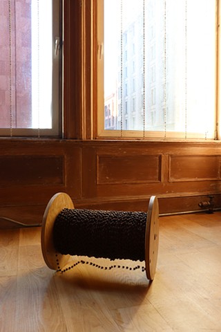 semilla santa, beaded curtain of coffee beans assembled onto chains , each about 8 inches apart, hang in front of two tall windows surrounded by a warm wood frame and wall panel below. EXTRAHERE, a wood cable reel wrapped tight with a continuous string of coffee beans rests on light wood floors. The golden glow through the windows illuminates the work and a small string of coffee beans trailing onto the floor. 