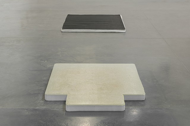 A close up of the two flat floor works lined up in the center of a dark gray museum floor. Reflecting Pool is in the foreground with the lip of the office chair mat facing towards us. The mattress topper underneath is cut to the dimensions of the mat and 