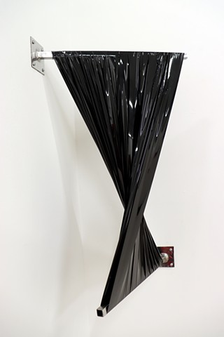 Strips of black shiny material wrap across two silver metal rods which extend outwards from a white wall and are anchored with square flanges. The piece is installed in a corner and the twist of the material creates a bow tie shape. The form shifts depend