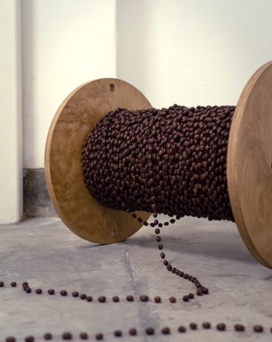 A wooden cable reel, which looks like an oversized spool, sits horizontally on it's side on a concrete floor in front of a white wall. The cylindrical middle of the cable reel is fully covered by layers of coffee beans, which have been glued together in p