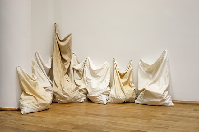 Eight pillowcases, stained from use, sit vertically huddled together on a wood floor in a corner by a column. They lean against the white museum walls reaching upwards. One pillowcase, intended for a body pillow, reaches the highest. They are all filled w