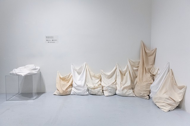 10 pillowcases, stained from use, sit vertically huddled together, overlapped and lined up on a gray floor in a corner against a white wall, all reaching upwards. The pillowcases are different shades of white, beige, and cream. One pillowcase, intended fo
