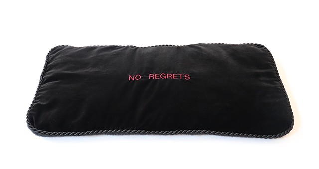 A black velvet lap pad sits on a white background. It’s rectangular, an inch and a half thick, and a twisted cord trim wraps around its edges. In the center of the lap pad, in small red machine embroidery, it reads “NO REGRETS” in all caps. If you look cl