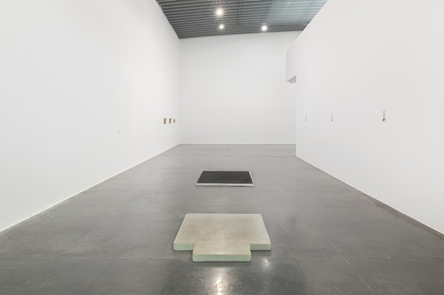 In a large museum with white walls and a high ceiling, two flat floor works are lined up in the center of a dark gray floor. %Reflecting Pool% is in the foreground with the lip of the office chair mat facing towards us. The mattress topper underneath is cut to the dimensions of the plastic mat and is blue turned pale seafoam green from use. %At Work (In Protest and in Care)% lies a few yards behind. It's used memory foam mattress topper is covered by a white fabric cover and placed underneath black diamond plate rubber flooring which has been cut to fit the topper's dimensions.