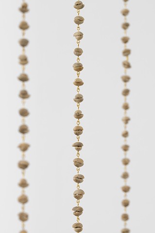 A detail shot of three unroasted coffee beans chains, vertically hanging in front of a white wall. The beans or rather seeds are a pale green jade-like color and have been paired together so that they created a rounded shape like beads. Reminiscent of a rosary or prayer beads, each bean has been drilled through and the pairs connected by small loops of golden brass wire.