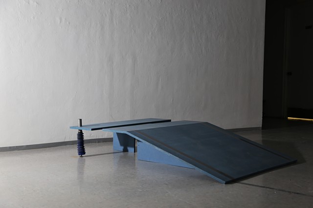A powder blue ramp and companion bench are positioned at a 45 degree angle leading up to a wall. The bench has a piston rod in lieu of foot, and peach-colored liquid puddles around the rod's blue rubber bootie. 