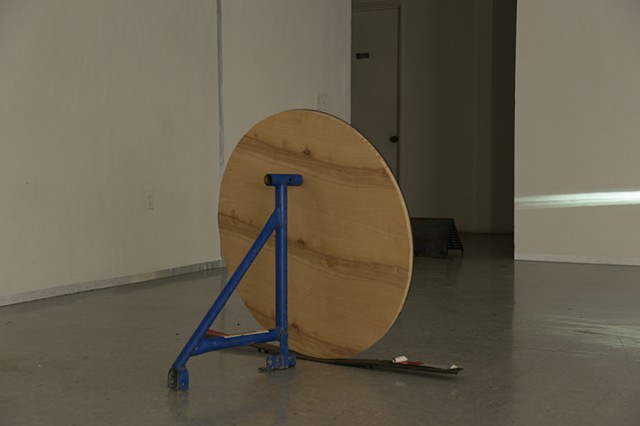 The sculpture "Rear View" is comprised of a 36-inch circle cut, wood-backed mirror resting against a 24-inch high ultramarine scaffold jack. The Mirror sits atop a 3-inch thin, 49-inch long rubber threshold strip positioned parallel to the mirror's length