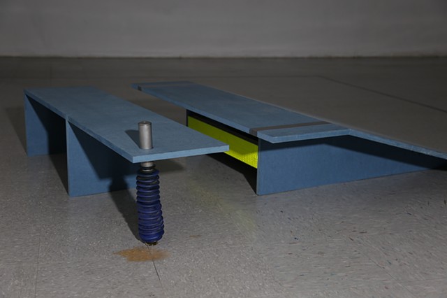 A powder blue ramp and companion bench are positioned at a 45 degree angle leading up to a wall. The bench has a piston rod in lieu of foot, and peach-colored liquid puddles around the rod's blue rubber bootie. 