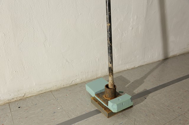 A 14-foot tall pipe with a square base leans against a wall, a blue-green biofoam is sandwiched between the scaffold base plates, leaving an impression, susceptible to foot traffic as it sits within the margins of a pathway.