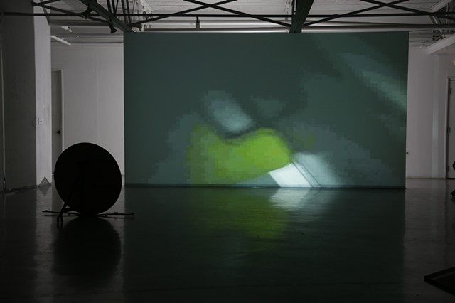 A freestanding wall with a projection of a bright white line at a 45-degree angle obstructed by a green shape on a black screen. The content of the screen's reflection is on the ground along with a silhouette of a freestanding circular sculpture.