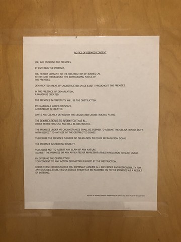 An 8.5-inch by 11-inch piece of paper is taped to the wall. Printed on the paper in capital letters reads the following text: NOTICE OF DEEMED CONSENT YOU ARE ENTERING THE PREMISES. BY ENTERING THE PREMISES, YOU HEREBY CONSENT TO THE OBSTRUCTION OF BODIES