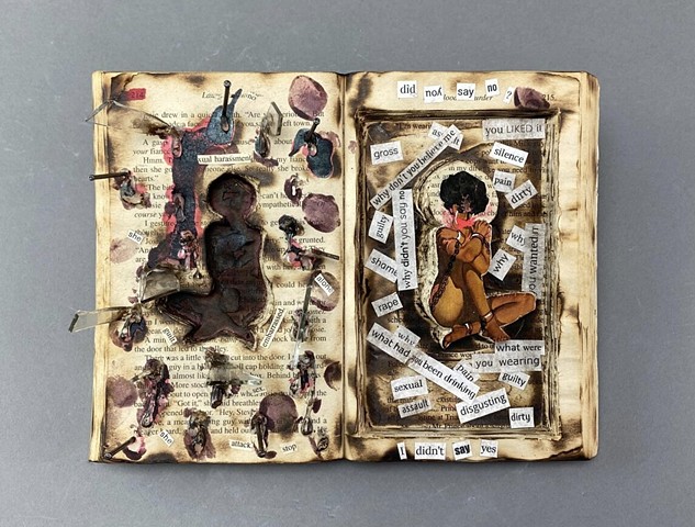 A small book which has been carved out to isolate the image of a woman sitting in chains. On the opposite side, only her silhouette is carved out and surrounded by nails and broken glass. The piece is covered in various words cut from printed paper.